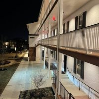 Visitation-hotel-exterior A two-story white brick building at night features a lit walkway, black shutters on windows, and white railings. External stairs lead to the upper floor. Leafless trees and landscaped areas border the walkway at Visitation Hotel Frederick MD.