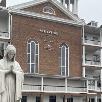 Visitation-hotel-frederick-exterior-front A white statue of the Virgin Mary stands with hands in prayer before a brick building labeled “Visitation Hotel Frederick” in Frederick, Maryland. The building has tall arched windows, white trim, and a small rooftop tower under an overcast sky.