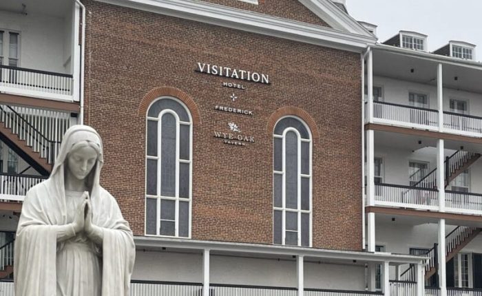 A white statue of the Virgin Mary stands with hands in prayer before a brick building labeled “Visitation Hotel Frederick” in Frederick, Maryland. The building has tall arched windows, white trim, and a small rooftop tower under an overcast sky.