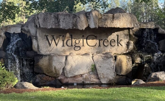 Widgi-creek-golf-course-bend-oregon A large stone sign reading Widgi Creek is set into a rock formation with small waterfalls on both sides. Green grass and trees surround this Bend Oregon Golf Course landmark, as sunlight casts shadows across the rocks.