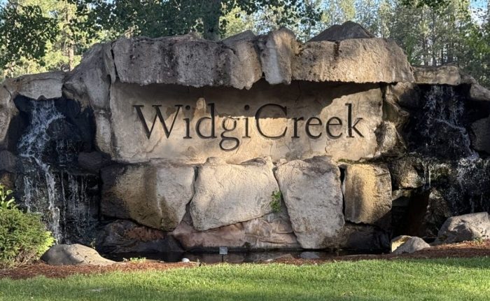 A large stone sign reading Widgi Creek is set into a rock formation with small waterfalls on both sides. Green grass and trees surround this Bend Oregon Golf Course landmark, as sunlight casts shadows across the rocks.